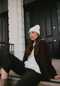 Modella seduta su una scalinata, indossa un berretto bianco lavorato a maglia, un maglione crema con orlo a coste e un cappotto scuro, abbinati a pantaloni neri larghi.