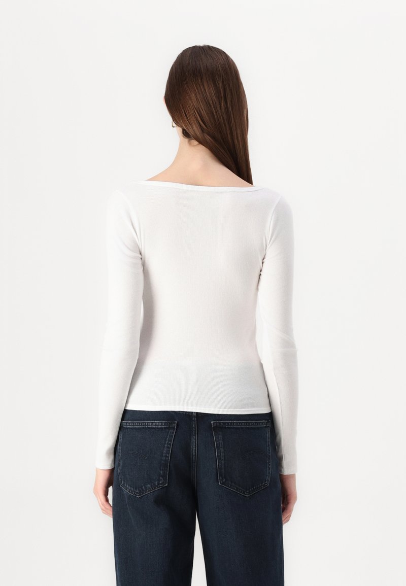 Weißes langärmliges, enganliegendes Oberteil mit quadratischem Ausschnitt, geripptem Textur und glattem Stoff, kombiniert mit dunkelblauen hochtaillierten Jeans.