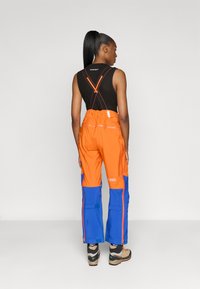 Abbigliamento outdoor colorato con parte superiore arancione e parte inferiore blu, spalline regolabili e tasche funzionali. Tessuto testurizzato.