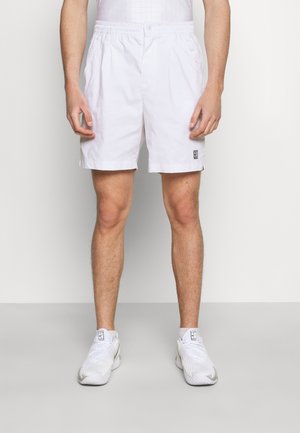 Homme portant un short de sport blanc avec taille élastique et des baskets blanches, debout contre un mur uni.