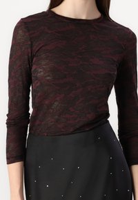 Model i en figursydd, genomskinlig bordeaux långärmad topp med ett strukturerat mönster, kombinerad med en högmidjad svart kjol prydd med små nitar.