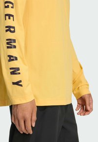 Geel shirt met lange mouwen en "GERMANY" in dikke zwarte letters op de linkermouw. Gemaakt van zachte stof, aansluitend ontwerp met een ronde hals.