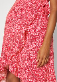 Vestido vermelho com padrão de manchas brancas, apresentando detalhe em babados ao longo da bainha. Tecido suave e silhueta em A, mostrado com a mão sobre o tecido.