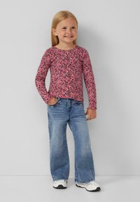 Maglietta rosa a maniche lunghe con motivo floreale, abbinata a jeans a gamba larga di colore blu chiaro. Il bambino sta in piedi con le mani nelle tasche, indossando scarpe bianche.