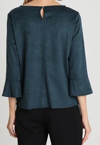 Blusa teal, strutturata, con maniche a tre quarti e polsini a campana. Dettaglio con apertura a chiave sul retro e vestibilità comoda. Design semplice ed elegante.