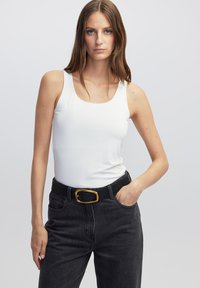 Witte mouwloze top met een ronde hals, gecombineerd met zwarte denimjeans en een zwarte riem met een goudkleurige ovale gesp.