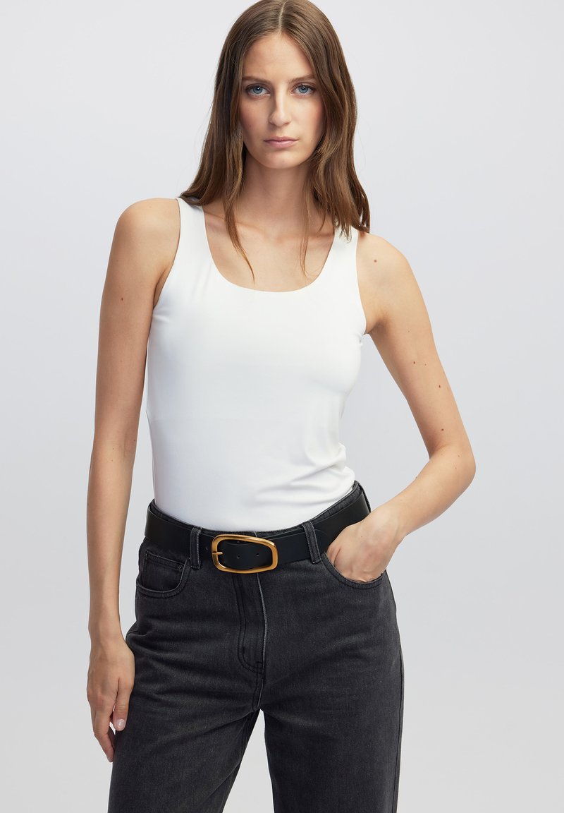 Witte mouwloze top met een ronde hals, gecombineerd met zwarte denimjeans en een zwarte riem met een goudkleurige ovale gesp.