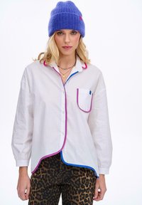 Bílá košile s růžovým a modrým lemováním, s kapesníkem. Kombinováno s kalhotami se vzorem leopardí kůže. Modrá pletená čepice na hlavě.