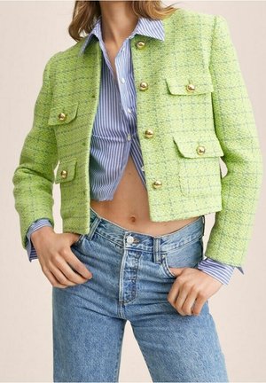 Frau trägt cropped limettengrüne strukturierte Jacke mit goldenen Knöpfen, blau gestreiftes Hemd und hoch taillierte blaue Jeans, Hände nahe den Taschen.