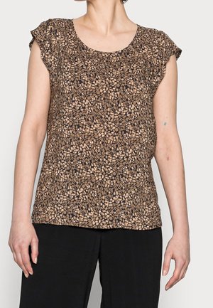 Kobieta ubrana w brązowo-czarną bluzkę z krótkim rękawem w wzory, z okrągłym dekoltem oraz czarne spodnie, stojąca na jednolitym tle.