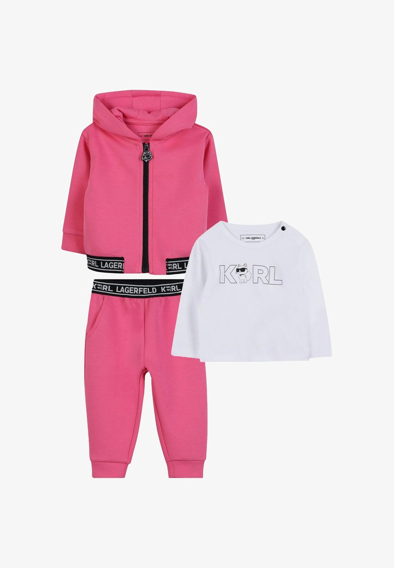 Roze zip-up hoodie en bijpassende joggers met zwarte accenten, gecombineerd met een witte langemouwen shirt met een grafische print.