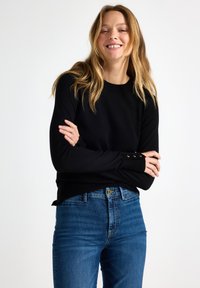 Zwarte gebreide trui met lange mouwen; heeft knopen op de manchetten. Gepaarde met lichtblauwe denim jeans in een slim fit.