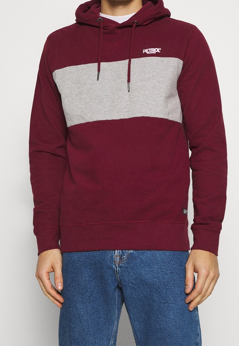 Hombre vistiendo una sudadera con capucha en colores burdeos y gris con el logo de "Petrol Industries", combinada con jeans de mezclilla azules, de pie contra un fondo blanco.