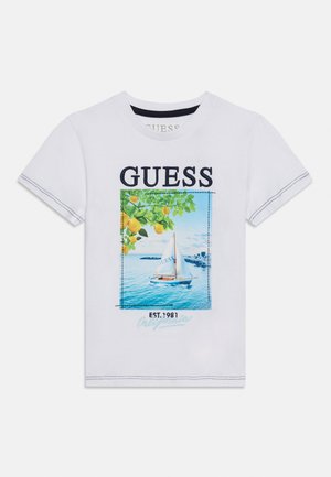 Hvid t-shirt med korte ærmer med teksten "GUESS" over en sejlbåd på blåt vand, en citrongren ovenover og "EST. 1981" under billedet.