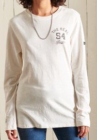 Långärmad, krämfärgad t-shirt i mjukt material, med en tryckt logotyp i grått på bröstet och en liten märkesetikett på ärmen.