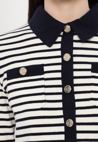 Striped knit top featuring navy and white colors, a collar, and six decorative gold buttons along the front, with two chest pockets.