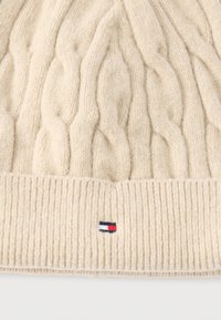 Kremfarvet strikket bæger med kabelmønster og ribkant, med et lille rektangulært logo i rødt, hvidt og marineblåt midt for.