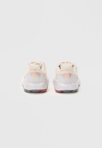 Une paire de chaussures de sport beiges vues de derrière, avec des semelles blanches et des accents rouges et gris sur les talons.