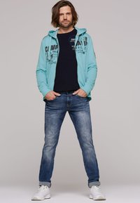 Light teal zip-up hoodie with printed graphics, paired with a navy t-shirt, blue jeans, and white sneakers. Casual fit, relaxed design.