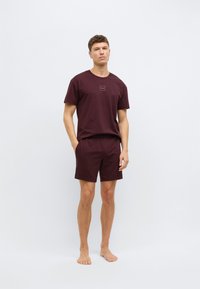 Tummanviininpunainen puuvillainen t-paita, jossa on pieni logo rinnassa, yhdistettynä vastaaviin shortseihin. Molemmissa on rento istuvuus ja sileä pinta.