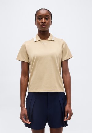 Kvinde iført beige kortærmet polo-shirt og marineblå plisserede shorts, stående mod en ensartet lys baggrund.