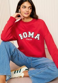 Felpa rossa con il testo "ROMA ITALIA", vestibilità comoda, abbinata a jeans a gamba ampia blu e sneakers bianche con strisce verdi.