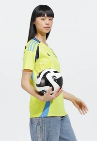 Svetlo rumena športna majica z modrimi poudarki in črnimi črtami, kombinirana s svetlo modrimi kavbojkami, drži črno-belo nogometno žogo.