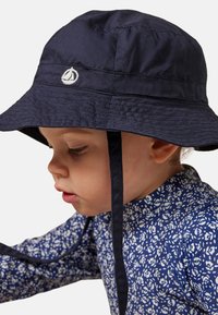 Marineblauwe emmerhoed met kinbandjes, met een wit zeilbootlogo. Wordt gedragen door een kind, gecombineerd met een blauw shirt met een bloemmotief en lange mouwen.