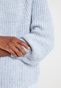 Hellblau gestrickter Pullover mit strukturiertem Muster, aufgerolltem Bund und geripptem Saum, mit einer Nahaufnahme einer Hand, die auf dem Ärmel ruht.