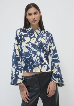 Eine Frau mit langen, dunklen Haaren, die ein blau-weißes, floral gemustertes Hemd mit Knöpfen und schwarze Lederhosen trägt. Sie zeigt ein Rosen-Tattoo auf ihrer linken Hand.