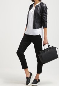 Giubbotto bomber in pelle nera sopra una camicia bianca con bottoni, pantaloni neri alla caviglia e scarpe slip-on nere. Borsa nera con doppi manici.