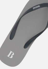 Pelēki flip-flopi ar teksturētu zolīti un melnām gumijas siksnām. Uz augšējās virsmas ir izcelta balta burta "B".