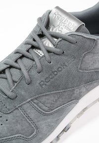 Gros plan sur une sneaker Reebok Classic en suede gris avec des lacets gris et une étiquette en métal argenté sur un fond blanc.