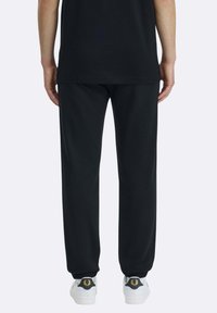 Schwarze Jogginghose aus weichem Stoff mit lockerem Schnitt und elastischen Bündchen. Kombiniert mit weißen Turnschuhen mit goldenem Emblem.