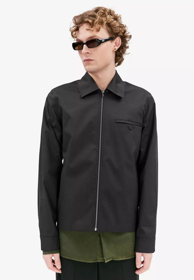 Zwarte zip-up jas met een kraag, details van een voorkwv pocket en lange mouwen, gelaagd over een groene shirt met een gladde textuur.