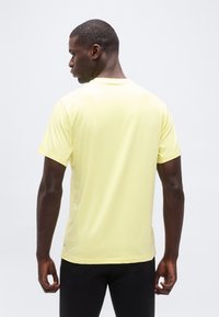 Άνδρας με πλάτη προς τα εμπρός, φοράει ανοιχτό κίτρινο κοντομάνικο πουκάμισο και μαύρο παντελόνι μπροστά σε απλό, φωτεινό φόντο.