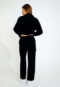 Schwarze Cordjacke im Cropped-Stil und passende Hose mit elastischem Bund. Das Outfit hat eine weiche Textur und wird mit weißen Sneakern kombiniert.