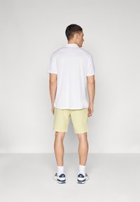 Balts īsām piedurknēm polo krekls, kas saskaņots ar gaiši dzeltenām šortiem. Modelis valkā baltas sporta kurpes ar zilām akcents un baltas zeķes.