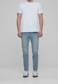 Valkoinen puuvillapaita, vaaleansiniset slim-fit-farkut, valkoiset ja harmaat lenkkarit mustilla pohjilla, seisomassa yksinkertaisella vaalealla taustalla.