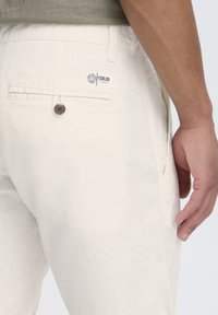 Pantalones de lino beige con un bolsillo trasero que cuenta con una etiqueta de logo y un botón marrón. La tela tiene una textura suave y un corte relajado.