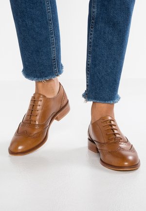 Brązowe skórzane buty sznurowane z detalami brogue; zaokrąglony nosek, niski obcas, w zestawie z krótkimi niebieskimi spodniami dżinsowymi z postrzępionymi krawędziami.