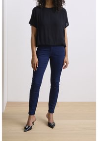 Haut noir à manches courtes ; jeans ajustés bleu marine ; mules à talons aiguilles noires avec détail de boucle ; tissu lisse ; coupe décontractée ; debout sur un sol en bois.