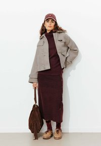 Manteau duffle en laine gris, pull côtelé bordeaux, jupe assortie avec des boutons sur le côté, sac à main en cuir brun avec des franges, et chaussures beiges à lacets.