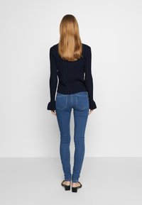 Top ajustado de manga larga con textura acanalada en color oscuro, combinado con jeans ajustados azul. Los jeans tienen un diseño entallado y bolsillos traseros, complementados con sandalias negras.