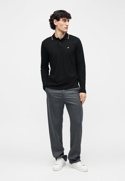 Άνδρας που φοράει μαύρο πόλο μακρυμάνικο, γκρι παντελόνι και λευκά αθλητικά παπούτσια, στέκεται με τα χέρια στις τσέπες, κοιτώντας προς το πλάι σε απλό φόντο.