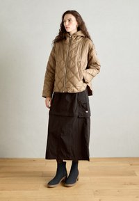 Veste à capuchon matelassée marron avec fermeture éclair, associée à une longue jupe noire dotée de poches, et des bottes noires mi-mollet avec une semelle en bois.