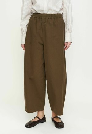 Persona indossa pantaloni larghi marroni con vita elastica, camicia a righe color crema e ballerine nere, in piedi su sfondo bianco.