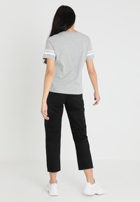 Mujer de pie de espaldas, vistiendo una camiseta corta gris con rayas blancas en las mangas, pantalones negros y zapatillas blancas, sobre un fondo blanco.