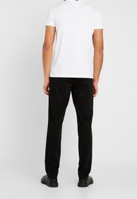 Muž stojící zády k nám, oblečený v bílém polo triku s krátkým rukávem, černých kalhotách a černých botách na jednotném pozadí.