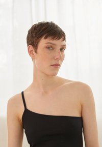 Top de malha preto com uma alça fina, apresentando um design justo e uma textura suave. A modelo tem cabelo curto e olhos verdes.